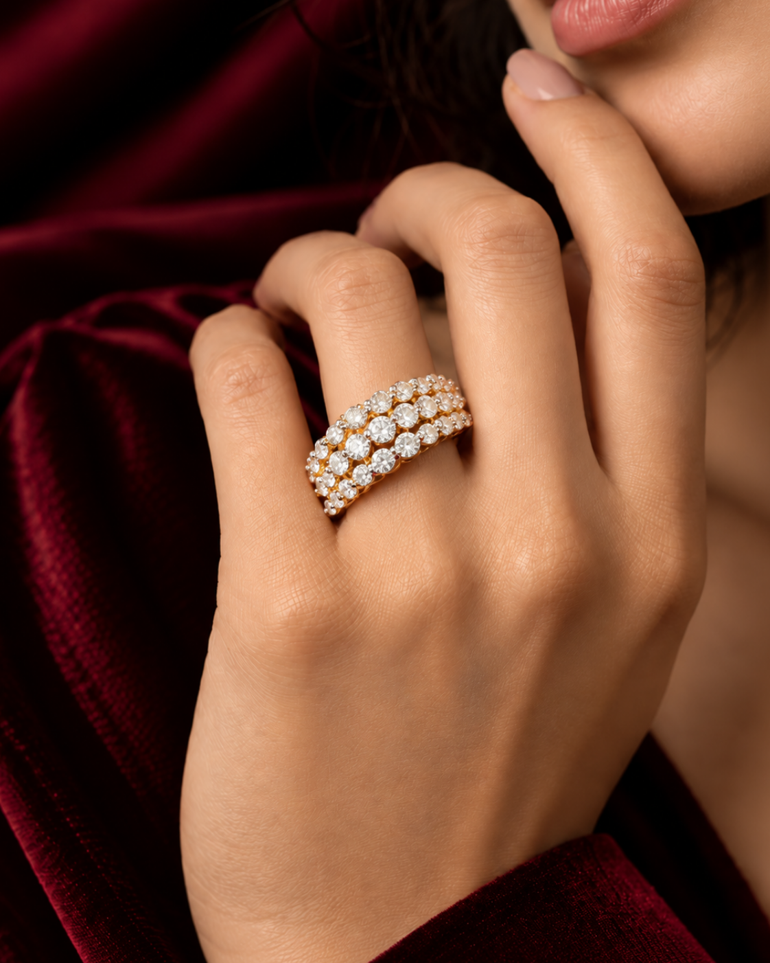 A close-up of a model's hand wearing a triple-row diamond eternity ring in yellow gold, set against a luxurious dark red velvet background.