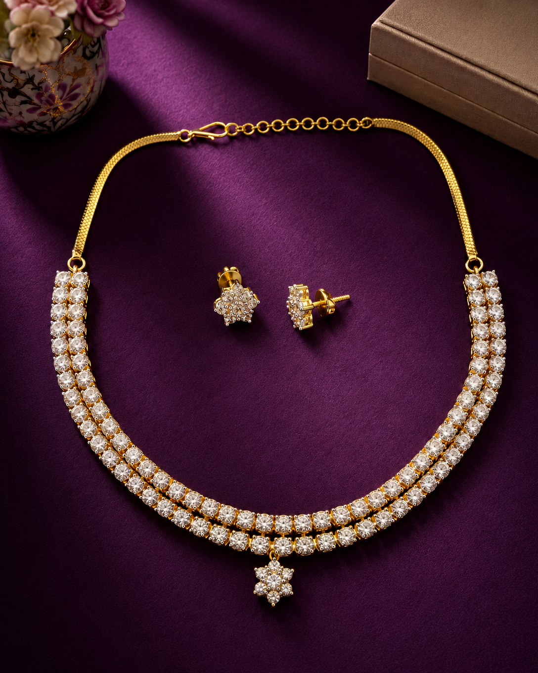 A gold-plated double-row necklace with matching stud earrings, both adorned with clear pavé stones and a central hanging flower motif, displayed on a deep purple satin fabric background.