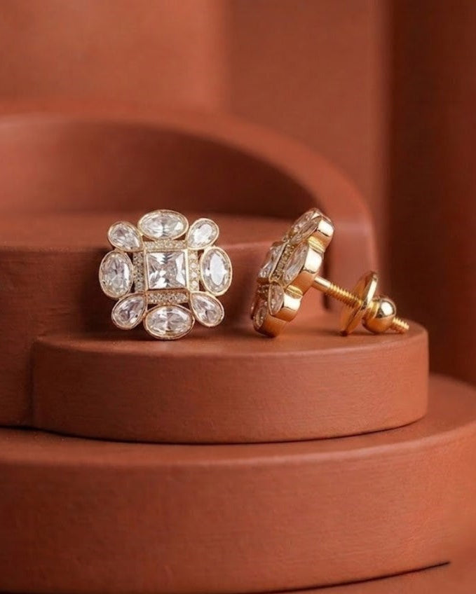 A pair of gold floral stud earrings with clear gemstones, displayed on a terracotta-colored tiered pedestal.