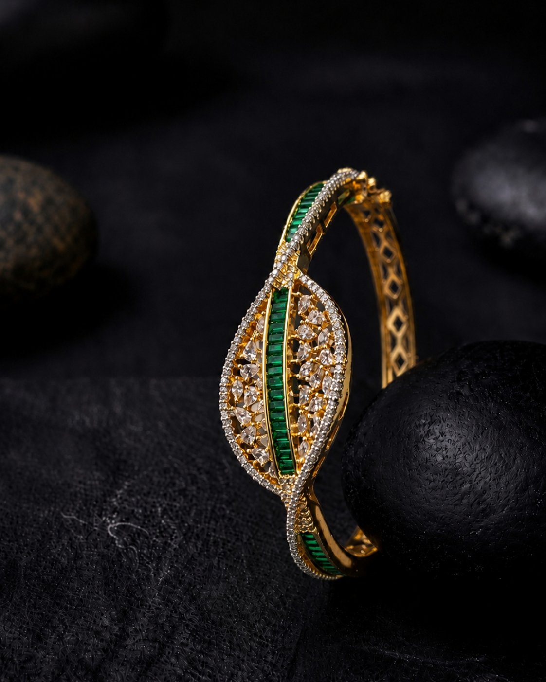A gold-toned bangle bracelet with a leaf-shaped design, featuring a central vertical column of emerald green stones and a surrounding pavé setting of small clear stones, displayed on a dark textured surface with black stones in the background.