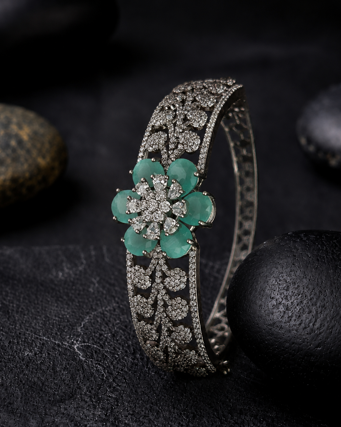 An ornate silver-toned hinged cuff bracelet with a central flower motif featuring green jade-like petals and pavé-set clear stones, displayed on a dark textured surface with black stones.