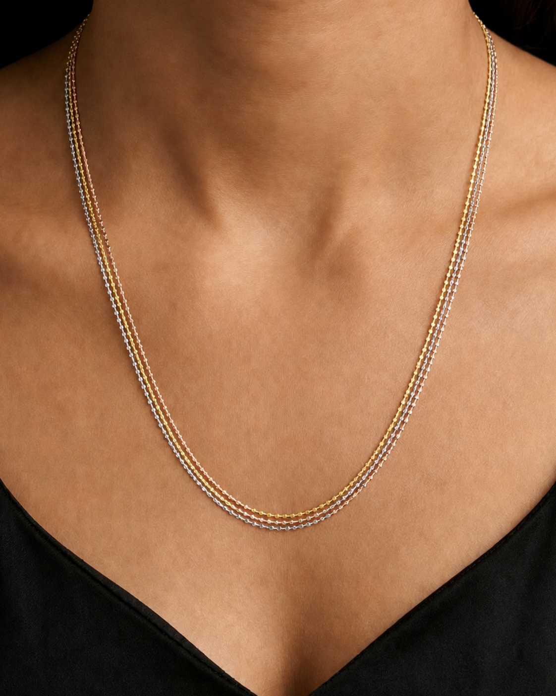 A close-up of a model's neck and upper chest wearing a delicate three-strand necklace with small gold and silver beads, paired with a black top.