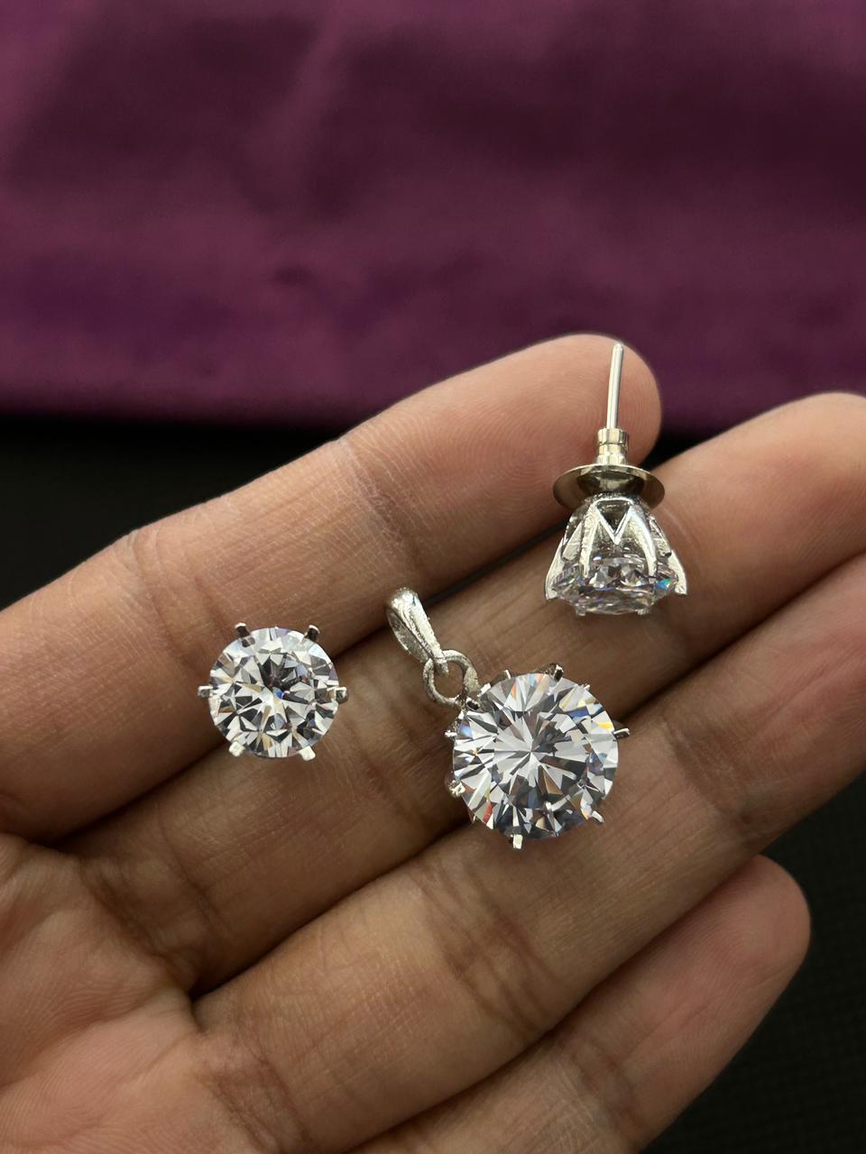 A pair of round-cut diamond stud earrings and matching pendant in silver settings, held in a person's hand against a dark background with a purple fabric surface behind.