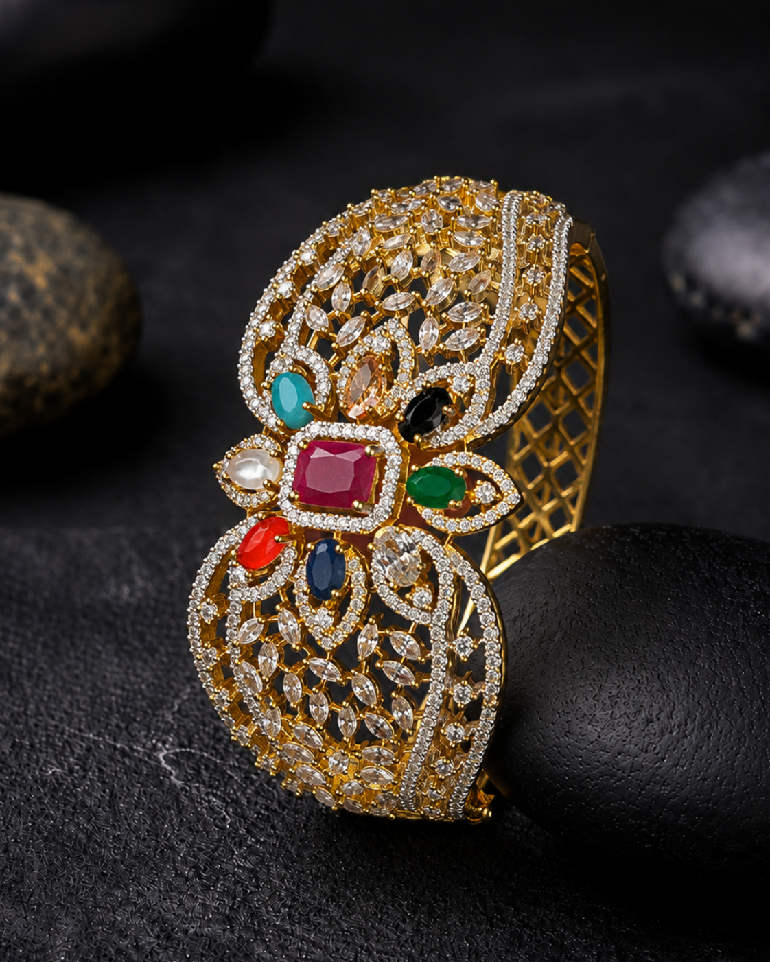 An ornate gold-toned cuff bracelet with intricate filigree detailing and colorful gemstone accents, displayed on a dark textured surface with smooth black stones.
