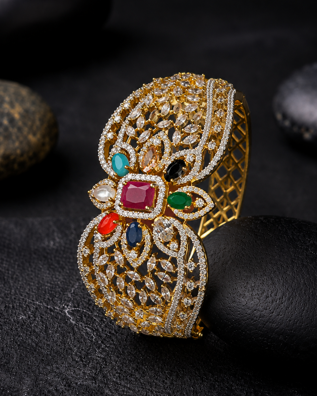 An ornate gold-toned cuff bracelet with intricate filigree detailing and colorful gemstone accents, displayed on a dark textured surface with smooth black stones.