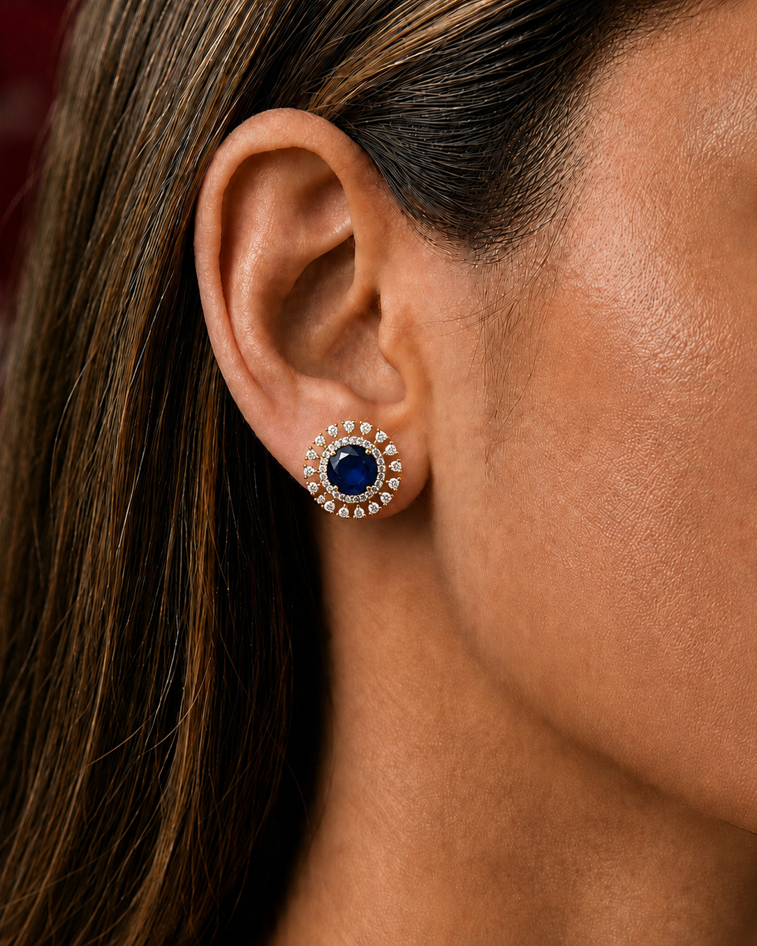 A close-up of a woman's ear wearing a single round stud earring with a deep blue center stone surrounded by a halo of small clear stones.