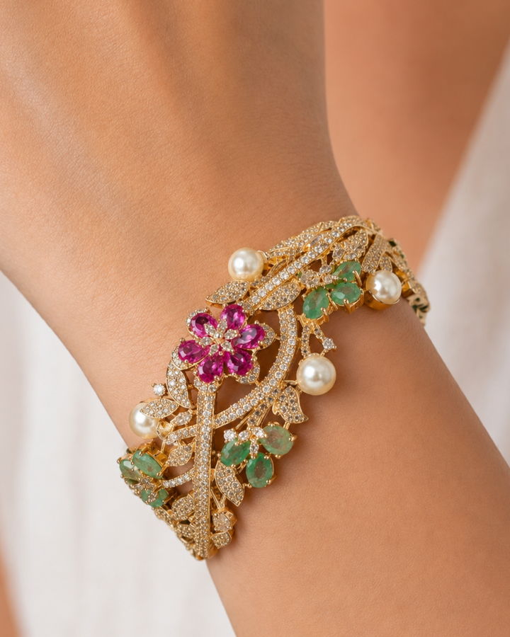 A close-up of a gold-toned bracelet worn on a wrist, decorated with a central pink flower, green leaves, and white pearl accents.