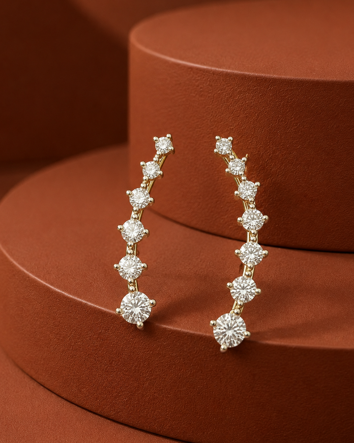 A pair of gold crescent-shaped dangle earrings with a row of clear round-cut stones, displayed on a textured terracotta-colored circular pedestal.