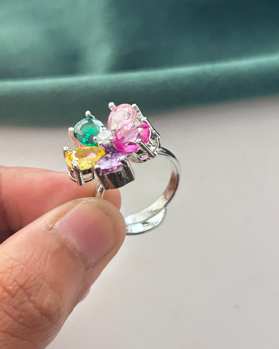 A silver-toned ring with a cluster of colorful gemstones including pink, green, yellow, and purple, held between fingers against a soft teal and white background.