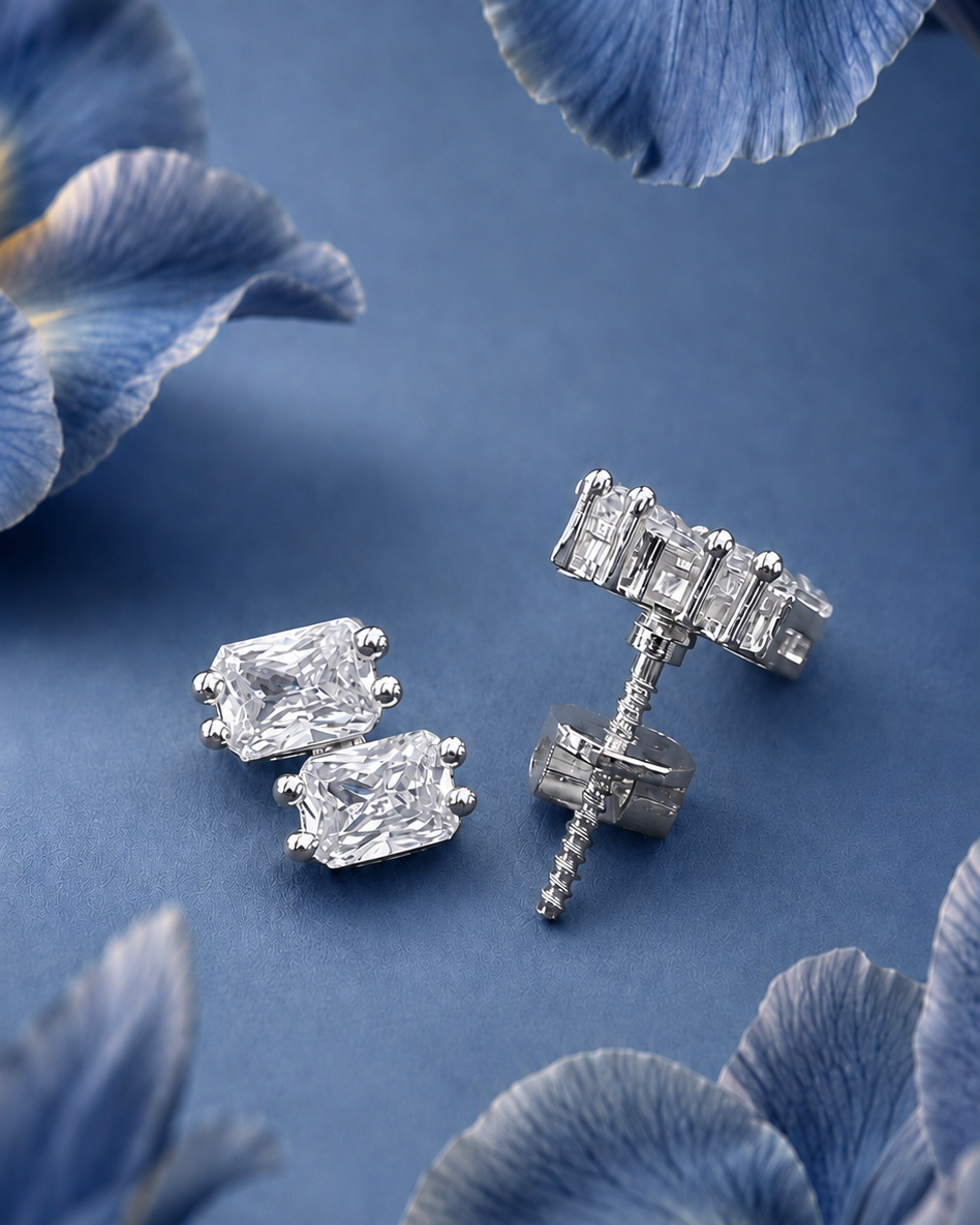 A pair of silver stud earrings with rectangular clear gemstones, one shown front-facing and the other angled to show the post and backing, set against a blue background with blurred blue flowers.