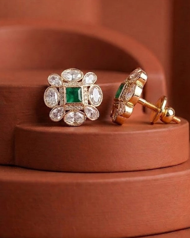 A pair of gold floral stud earrings with a central green square and clear surrounding stones, shown on a terracotta-colored display stand.