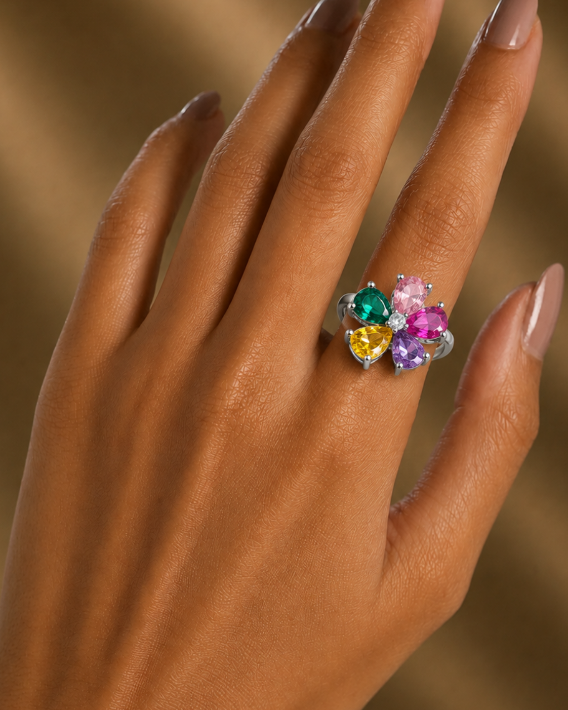 A close-up of a hand with manicured nails wearing a silver ring adorned with a cluster of colorful heart-shaped gemstones in green, yellow, and pink, set against a warm, softly blurred brown background.
