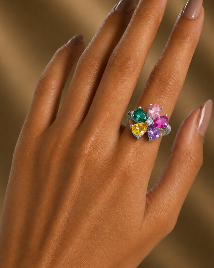 A close-up of a hand with manicured nails wearing a silver ring adorned with a cluster of colorful heart-shaped gemstones in green, yellow, and pink, set against a warm, softly blurred brown background.