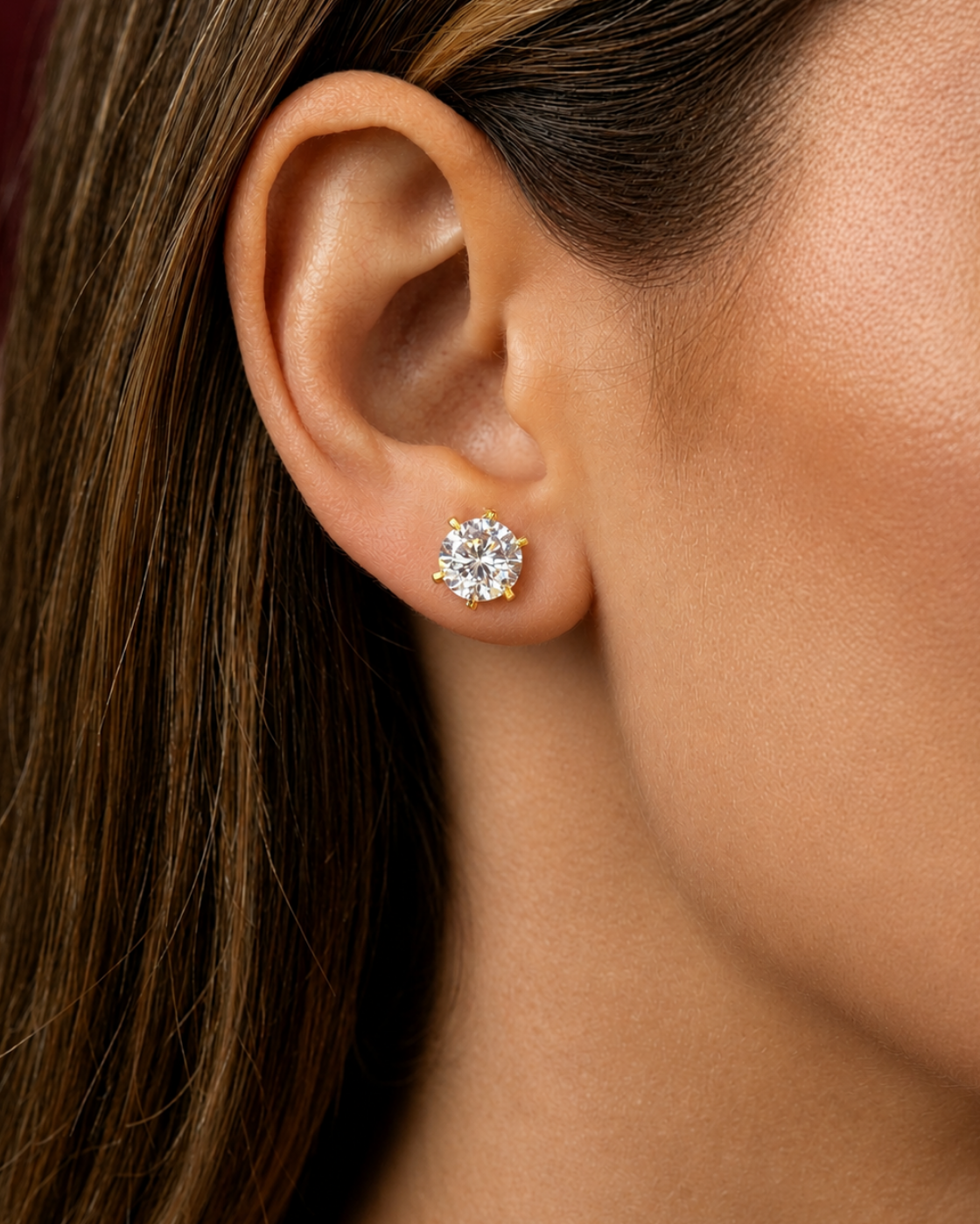 A close-up of a woman's ear wearing a small round brilliant-cut diamond stud earring in a yellow gold setting.
