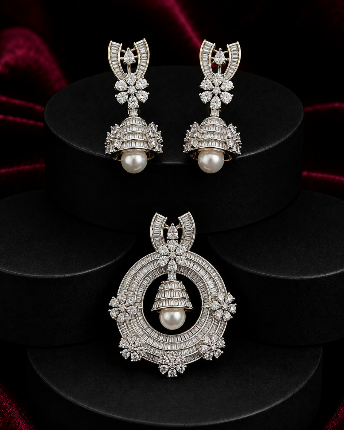 A pair of silver-toned Jhumka earrings and a matching Jhumka pendant displayed on black cylindrical pedestals against a dark red velvet background. The jewelry is adorned with pavé-set clear stones and a central white pearl.