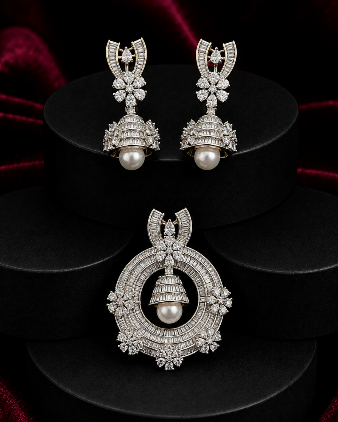 A pair of silver-toned Jhumka earrings and a matching Jhumka pendant displayed on black cylindrical pedestals against a dark red velvet background. The jewelry is adorned with pavé-set clear stones and a central white pearl.