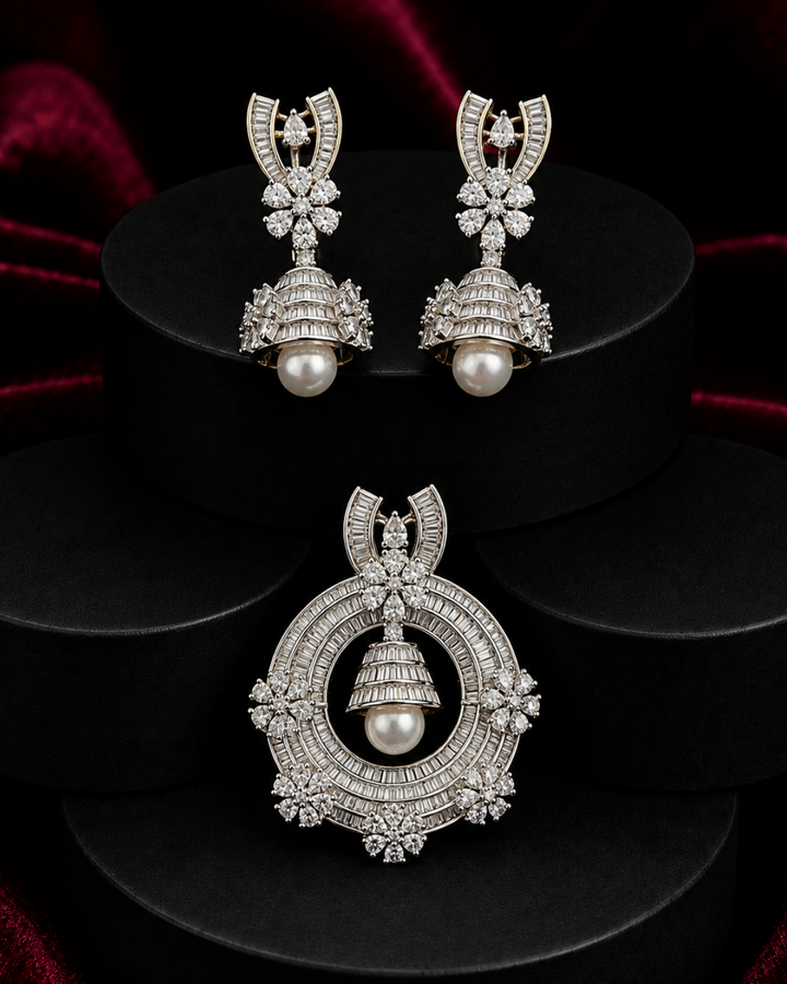 A pair of silver-toned Jhumka earrings and a matching Jhumka pendant displayed on black cylindrical pedestals against a dark red velvet background. The jewelry is adorned with pavé-set clear stones and a central white pearl.