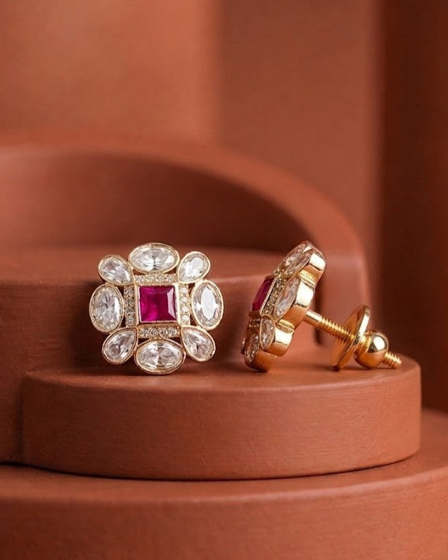 A pair of gold floral stud earrings with a central square pink gemstone and surrounding clear marquise and round stones, displayed on a terracotta-colored tiered pedestal.