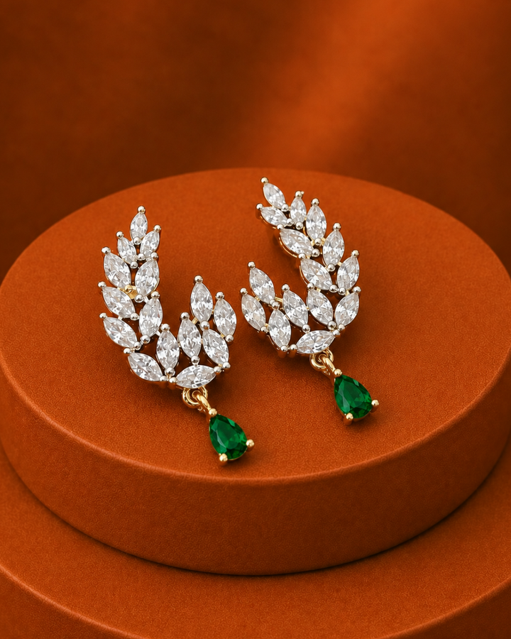A pair of gold leaf-shaped drop earrings adorned with marquise-cut clear stones and green teardrop emerald drops, displayed on a round brown pedestal against a matching brown background.