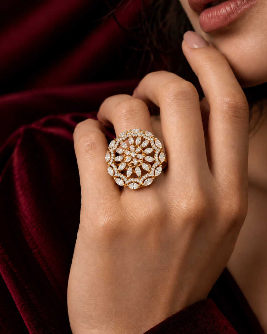 A close-up lifestyle shot of a woman's hand wearing a large, intricate circular ring with a floral mandala pattern of clear stones and warm yellow-gold accents, set against a dark red velvet background.