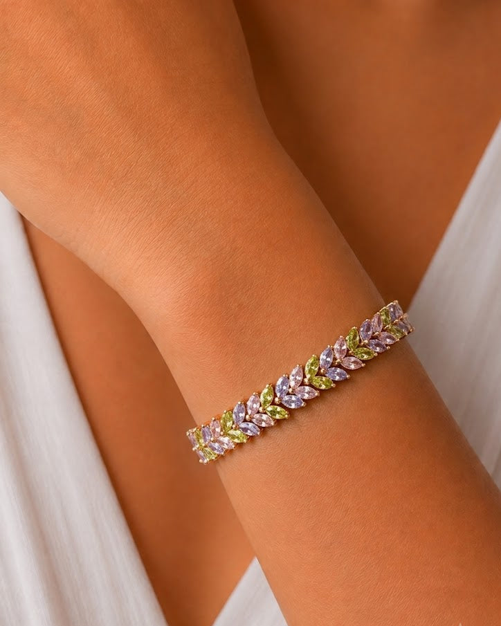 A close-up of a delicate bracelet worn on a woman's wrist, featuring a leaf-like pattern with light pink, lavender, and yellow-green stones in a gold-toned setting, against a white dress.
