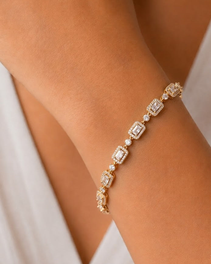 A close-up of a delicate gold bracelet with rectangular clear gemstones, worn on a person's wrist against a soft white garment background.