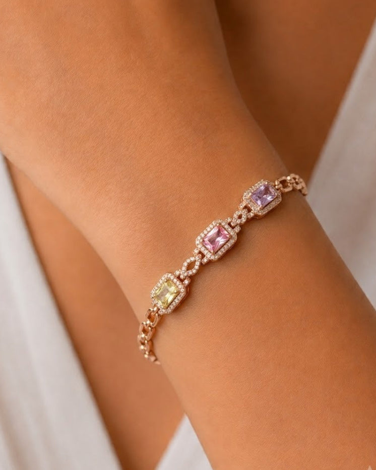 A close-up of a delicate rose gold bracelet with three colorful gemstone clusters (green, pink, and purple) worn on a woman's wrist.