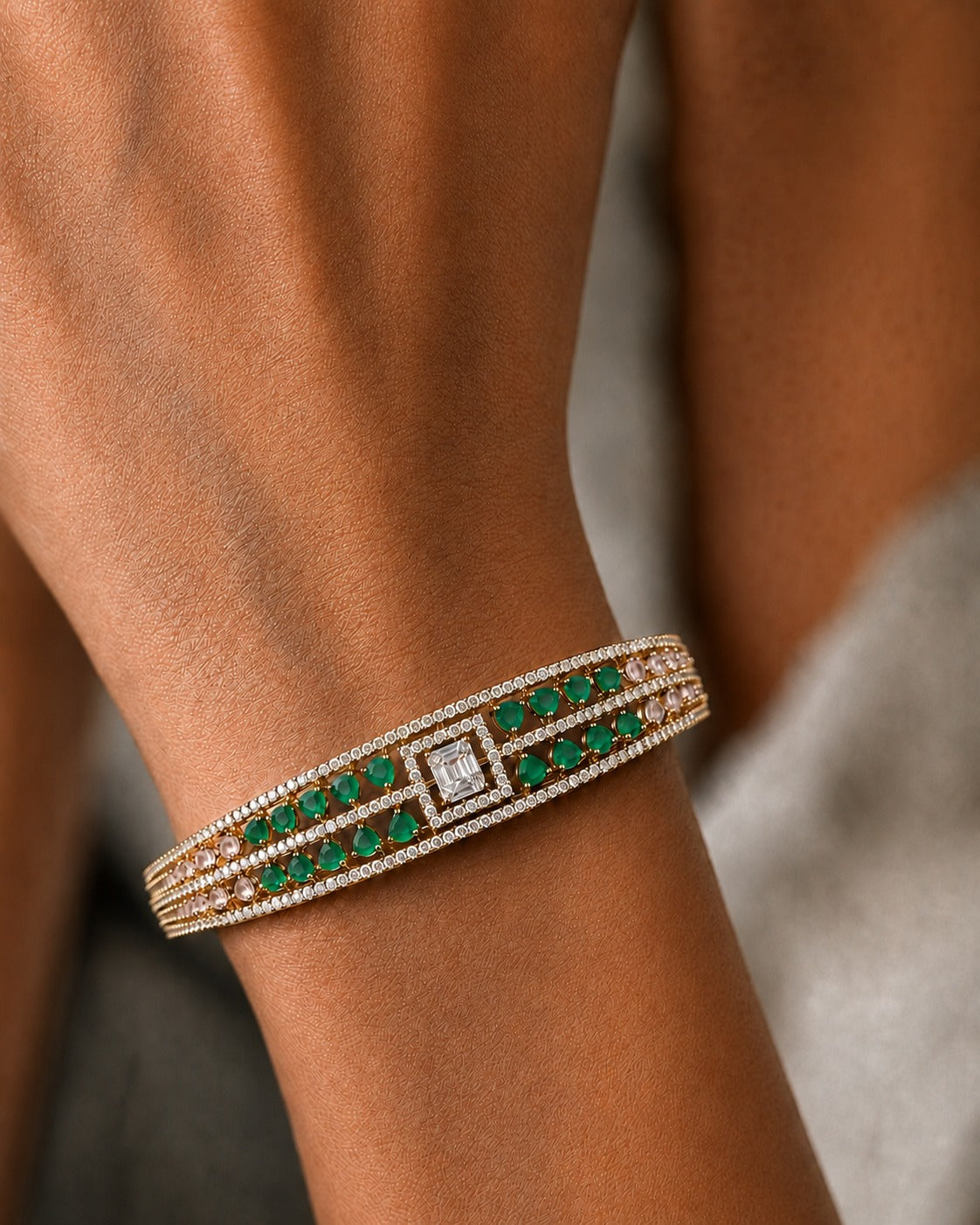 A close-up of a gold-toned cuff bracelet with green emerald-cut stones and a central diamond, worn on a person's wrist.