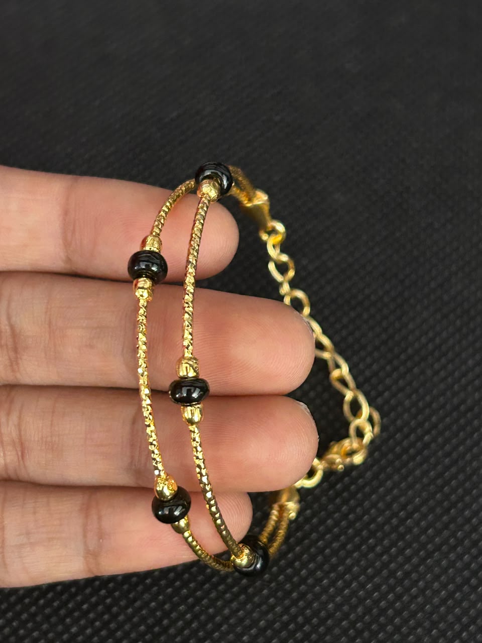 A close-up of a hand holding a gold-toned double-strand bracelet with small black beads and a chain link section, set against a dark textured background.