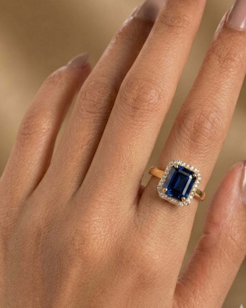 A close-up of a hand wearing a gold ring with a rectangular deep blue gemstone surrounded by small clear stones, set against a soft warm beige background.