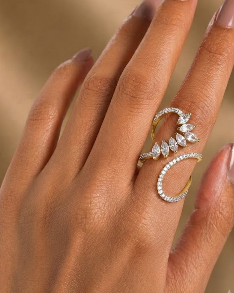 A close-up of a hand with manicured nails wearing a gold and diamond ring with a crescent and leaf design, set against a soft, warm beige background.