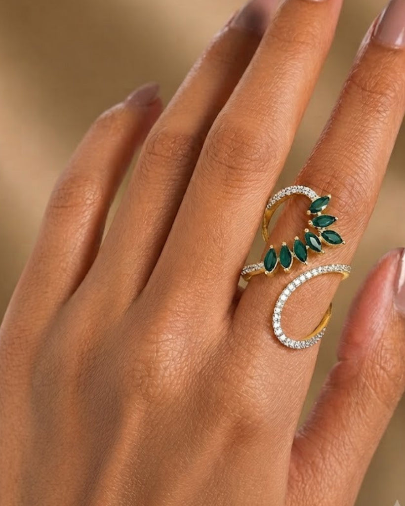 A close-up of a hand with manicured nails wearing a gold ring featuring a crescent-shaped design with green marquise-cut stones and a row of clear stones.