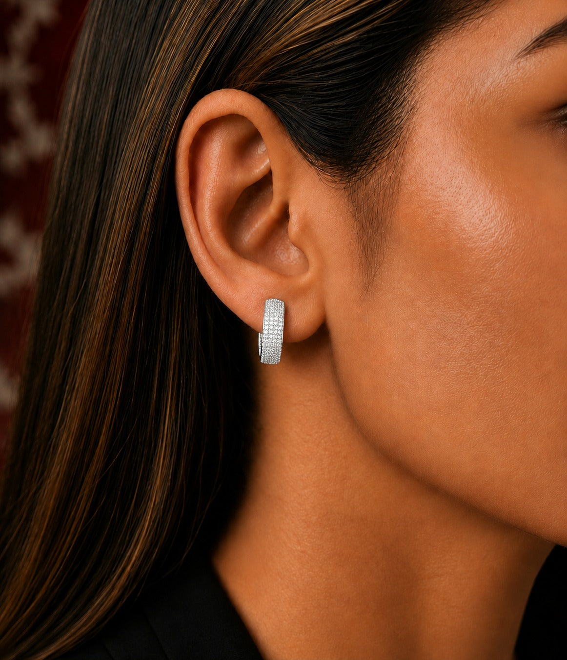 A close-up of a model's ear wearing a small silver hoop earring with a textured, pavé-set appearance.