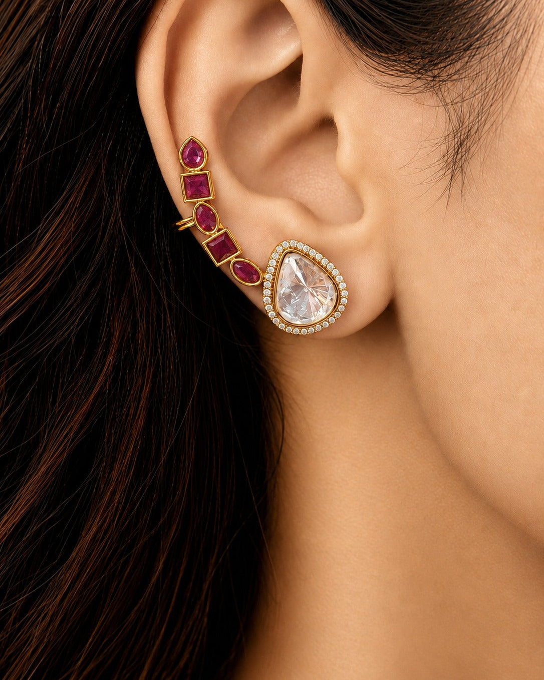 A close-up of a model's ear showcasing a gold ear cuff stack. The stack features a teardrop-shaped clear gemstone surrounded by a halo of small clear stones, with a vertical row of rectangular deep pinkish-red gemstones set in gold on the helix.