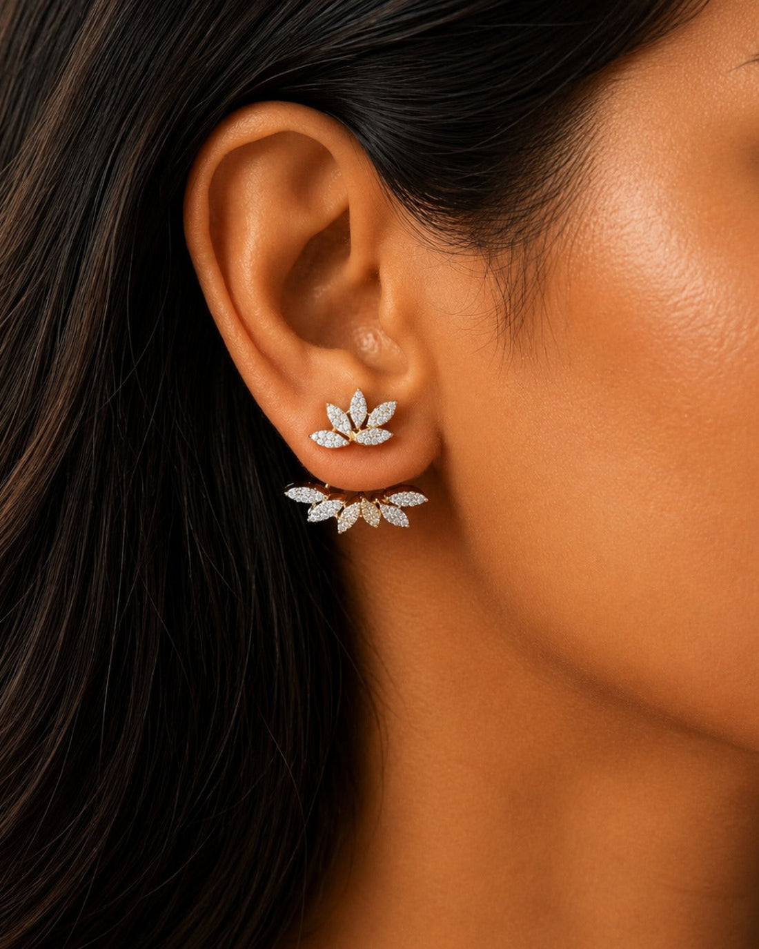 A close-up of a model's ear wearing a pair of silver-toned leaf-shaped stud earrings set with clear stones.