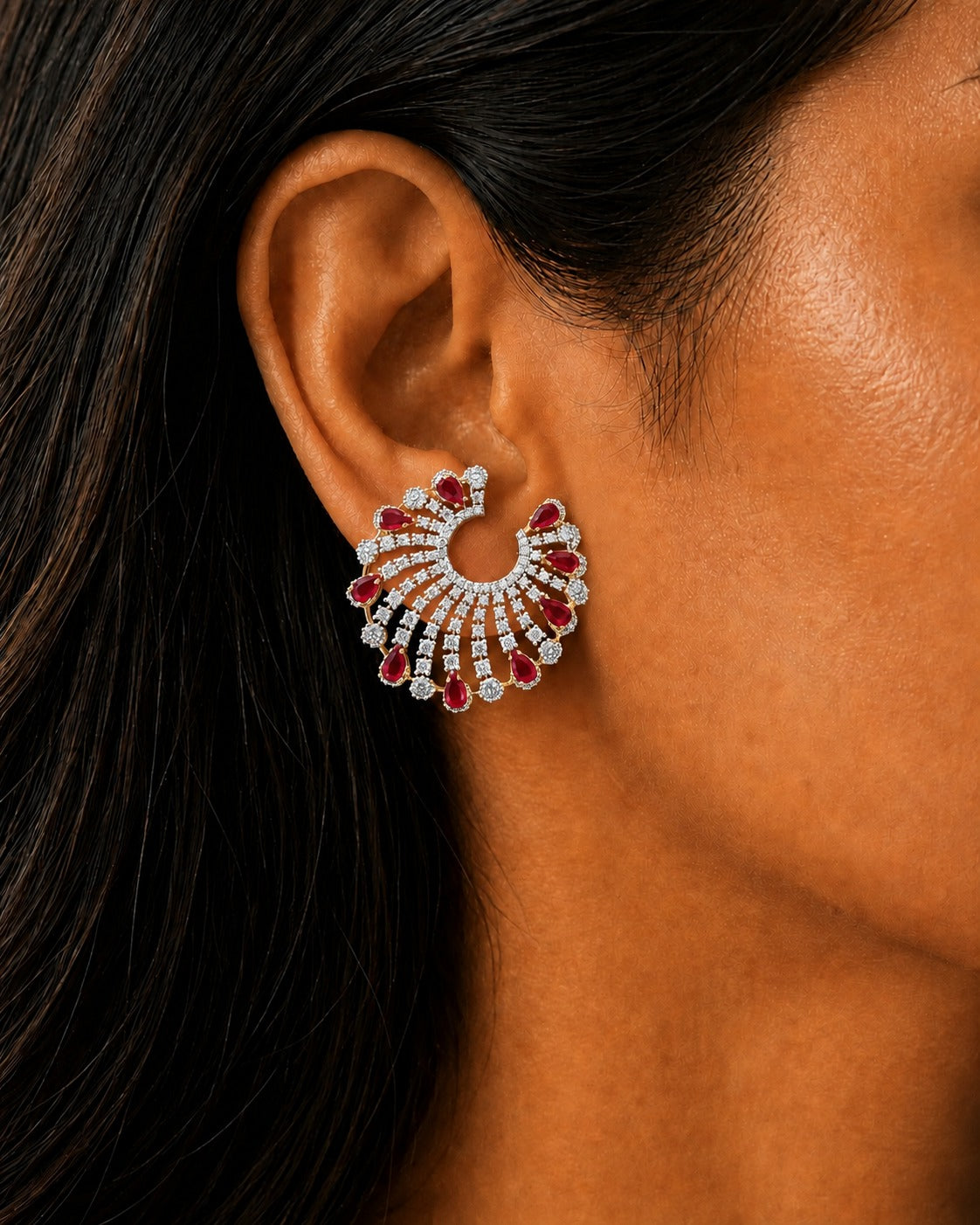 A close-up of a model's ear wearing a dramatic, multi-tiered crescent-shaped stud earring. The earring features silver-toned metal set with clear stones and deep red gemstones, photographed against the model's warm skin and dark hair.