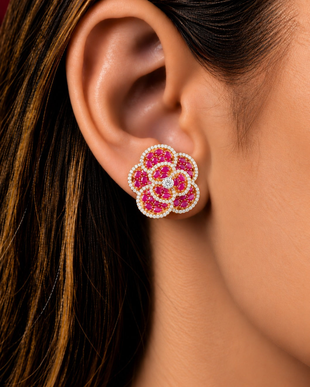 A close-up of a model's ear wearing a flower-shaped stud earring with bright pink gemstones and a border of small clear stones.