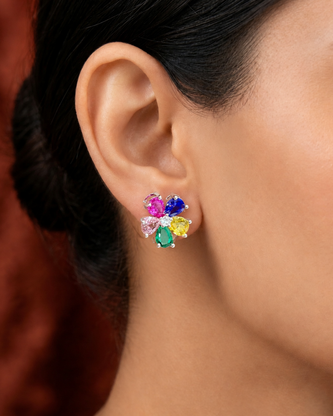 A close-up of a model's ear wearing a small cluster stud earring with five colorful gemstone petals in pink, blue, green, yellow, and clear stones on a silver-tone setting.