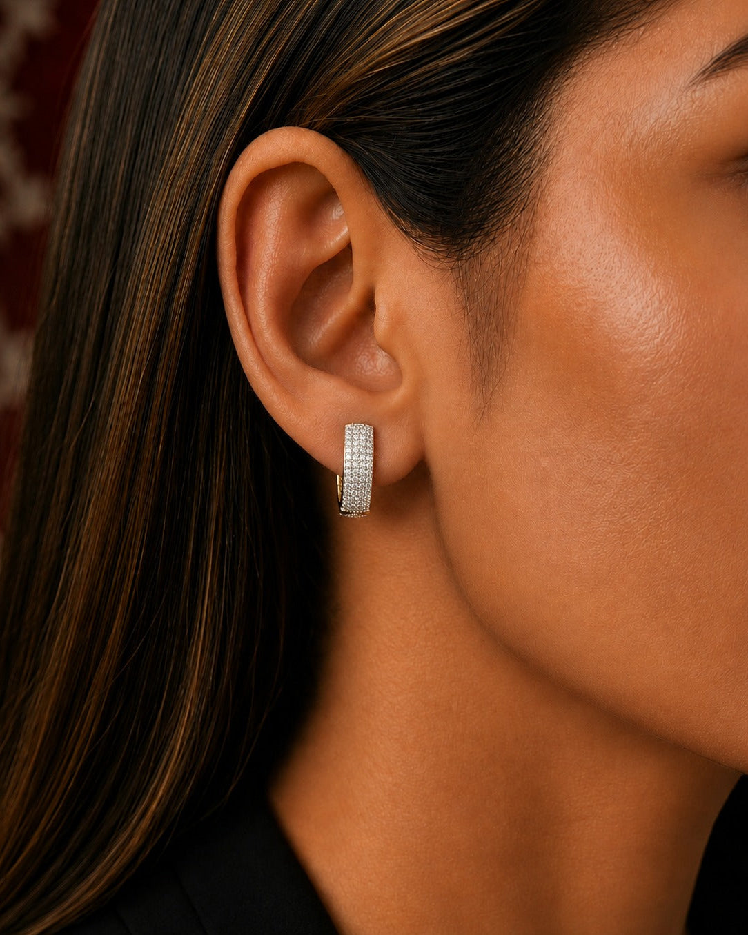 A close-up of a model's ear wearing a small silver hoop earring paved with clear stones.