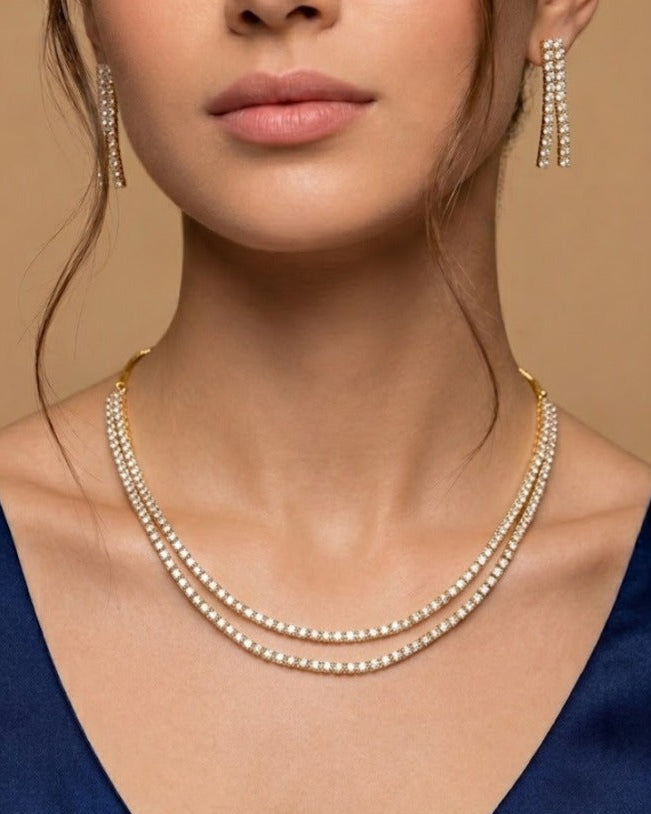 A close-up of a model wearing a double-strand gold tennis necklace with clear stones and matching drop earrings, paired with a dark blue satin top against a warm beige background.