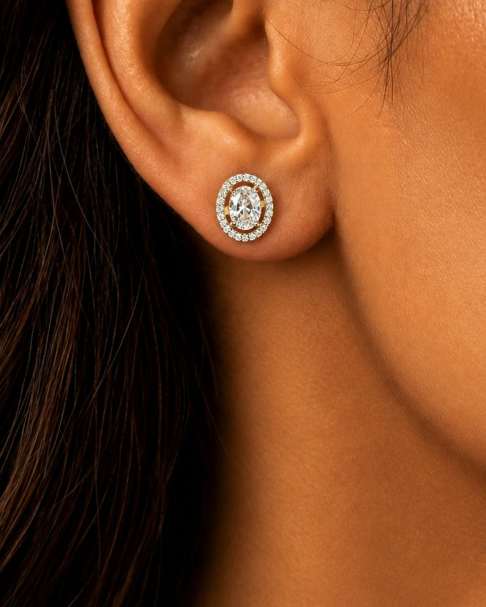 A close-up of a person's ear wearing a small oval stud earring with a silver-toned frame and clear stones.