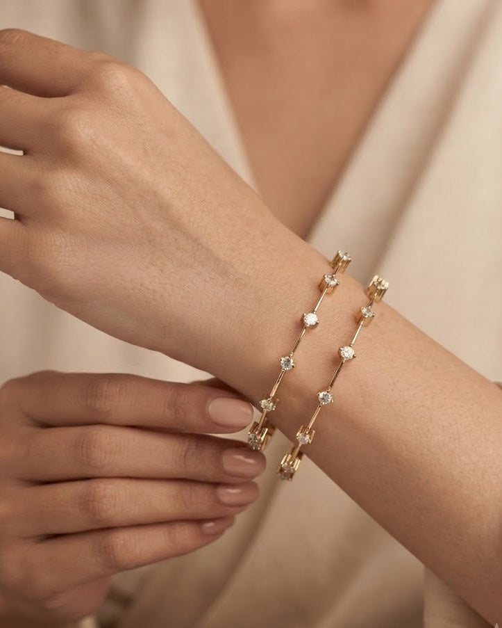 A close-up of a person's wrist wearing two delicate gold bracelets adorned with small clear stones, set against a soft beige robe.