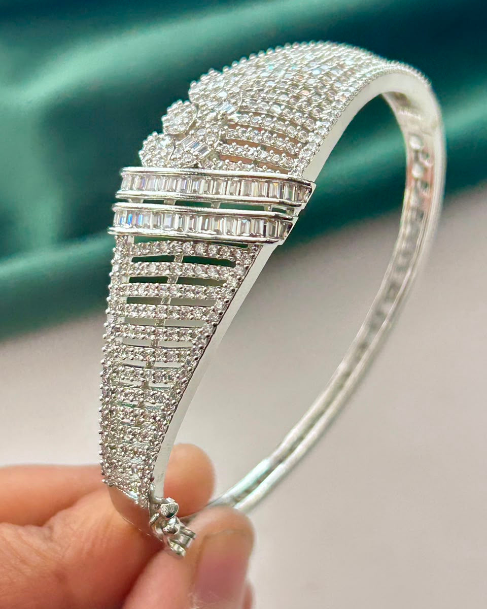 A close-up of a silver-toned bangle bracelet covered with clear pavé stones, held by a hand against a teal satin fabric background.