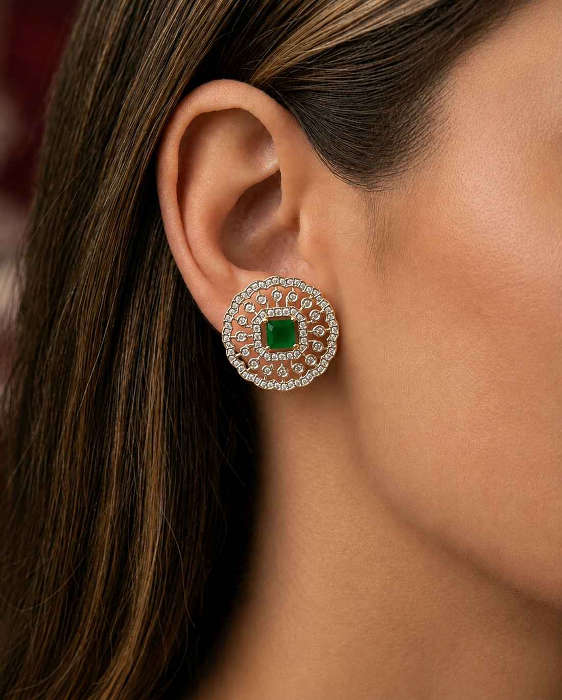 A close-up of a woman's ear wearing a circular stud earring with a central green square stone surrounded by a halo of small clear stones in a silver-toned setting.