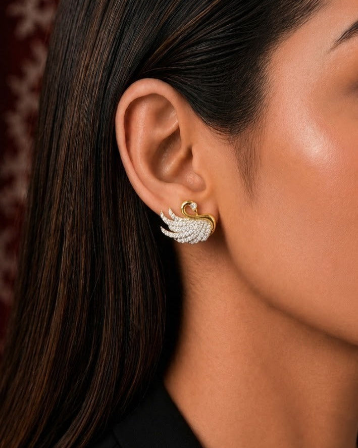 A close-up of a woman's ear wearing a gold and white pavé wing-shaped stud earring.