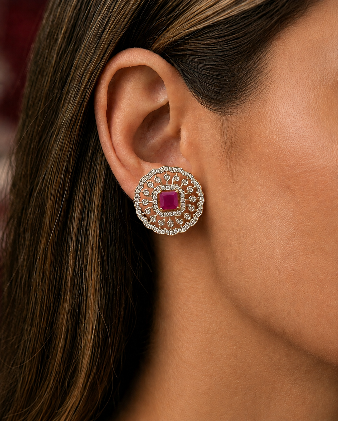 A close-up of a woman's ear wearing a circular stud earring with a central pinkish-red square gemstone surrounded by a halo of small clear stones.