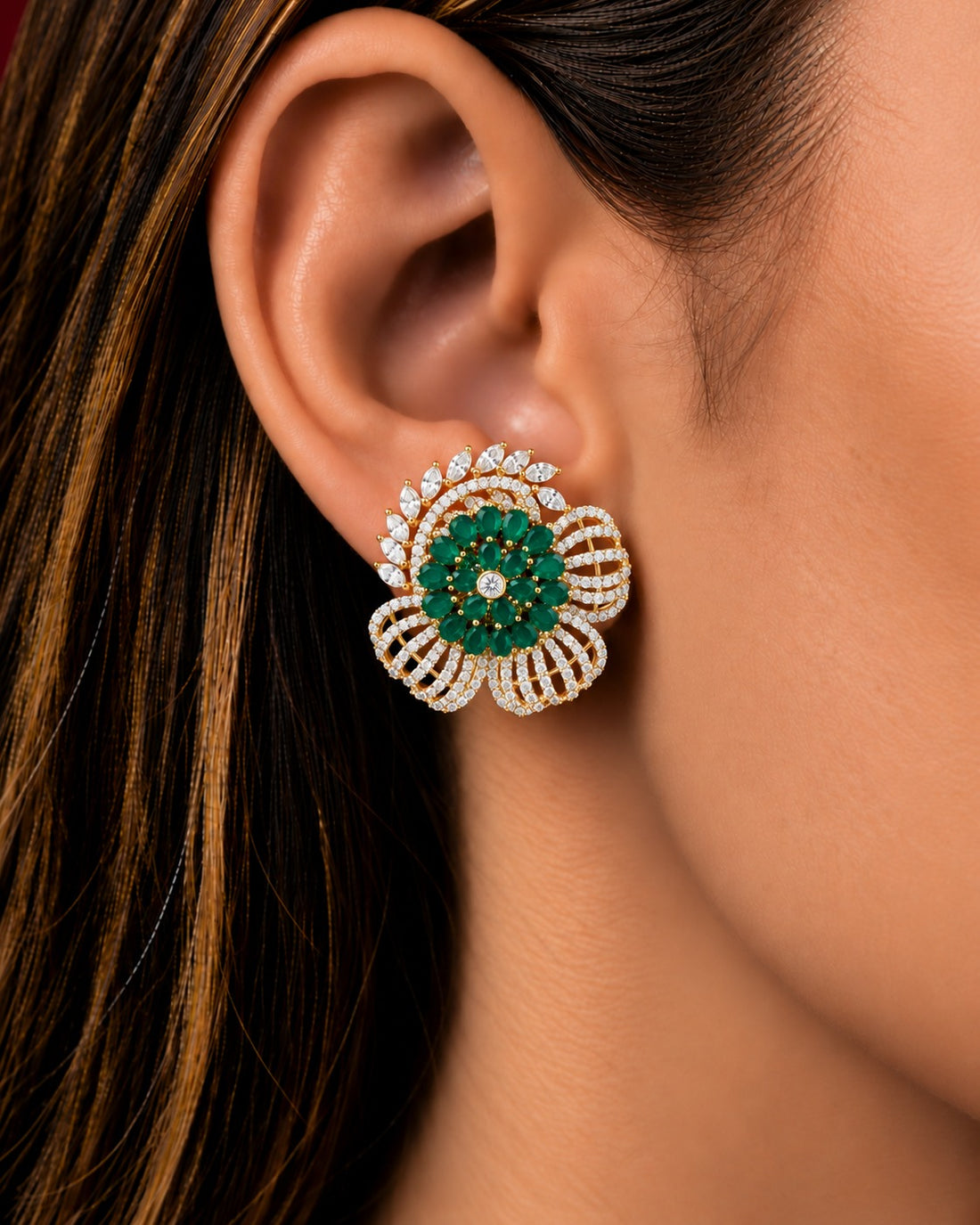 A close-up of a woman's ear wearing a detailed floral stud earring. The earring features a central green stone cluster surrounded by a gold-tone frame set with small clear stones, and layered petal-like sections on either side.