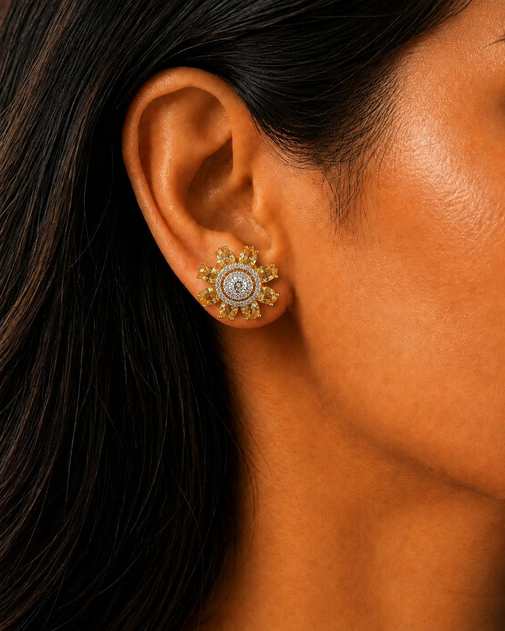 A close-up of a woman's ear wearing a gold-toned mandala-style stud earring with a clear central stone and surrounding small stones.
