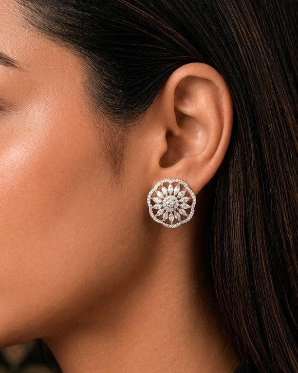 A close-up of a woman's ear wearing a round silver-toned stud earring with a detailed floral or sunburst pattern of small clear stones.