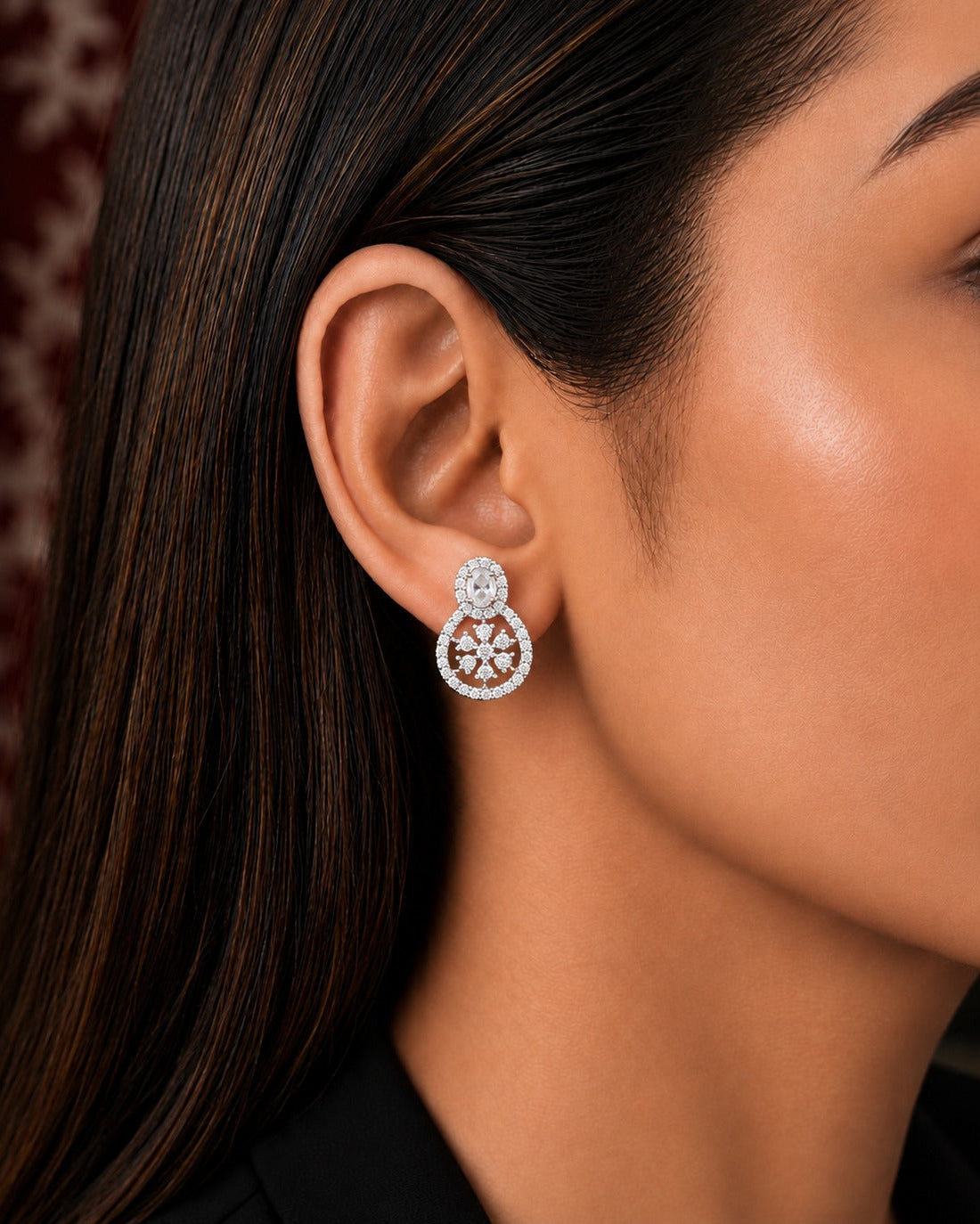 A close-up of a woman's ear wearing a silver-toned dangle earring with a circular halo of small clear stones and a central floral/starburst motif.